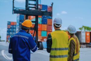 Construction workers in safety gear inspecting a shipping yard with stacked containers, representing supply chain challenges and tariff impacts on materials.